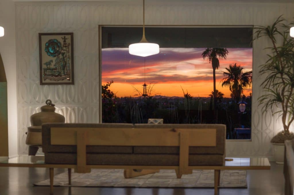 Rear view of a mid-century couch in a stylish living room, facing a massive window framing a dramatic tropical sunset with palm trees and glowing skies.